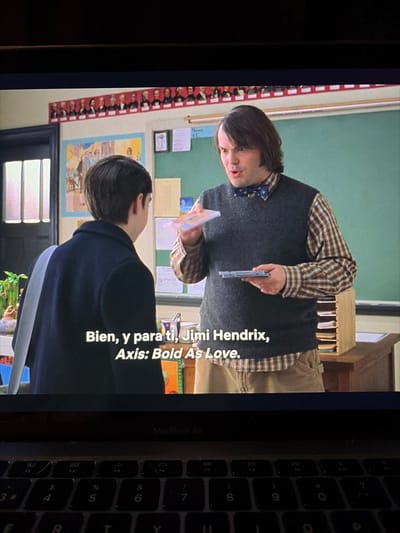 A man wearing a plaid shirt, dark gray sweater vest, and khakis stands in a classroom, holding two CDs. He's facing a person with dark hair, wearing a dark blue jacket with a light gray stripe. In the background is a green chalkboard, posters, and a row of photos. The text on the screen reads, "Bien, y para ti, Jimi Hendrix, Axis: Bold As Love." A MacBook Air keyboard is visible in the lower right corner.