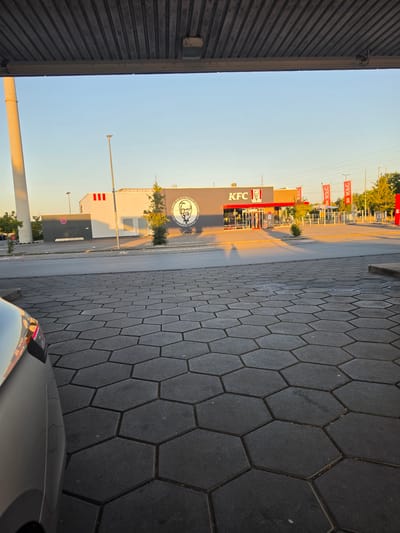 In Bischofsheim, Germany, a KFC restaurant is visible under a clear sky during what appears to be either sunrise or sunset, casting long shadows. The building features the iconic KFC logo and signage, with red and white accents. Red banners with the KFC logo are also displayed. In the foreground, a section of a silver car is visible, its reflection captured on its metallic surface. The ground is paved with dark gray hexagonal tiles. A tall white pillar is present on the left side of the image. The scene suggests an outdoor dining or drive-thru area associated with the fast-food establishment.