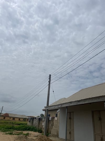 The image captures a scene in the city of Minna, Nigeria, under a cloudy sky. In the foreground, a concrete wall is visible, adorned with graffiti. Clothes are hanging on a clothesline attached to the wall. To the right, a building with a corrugated metal roof and light-colored walls has two doors. In the middle ground, more buildings with corrugated metal roofs are present, set against a backdrop of overgrown green grass and dirt. Power lines stretch across the sky, supported by wooden poles. The overall impression is one of a residential area in an urban or semi-urban setting.