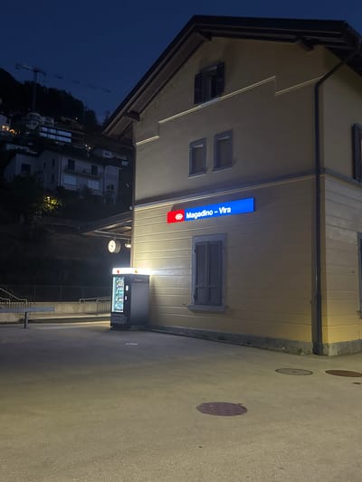 The image depicts the exterior of a train station in Magadino, Switzerland, at night. The station building is painted a light yellow and features a blue illuminated sign that reads "Magadino - Vira". Two small windows with dark shutters are visible on the upper facade, along with a larger window with similar shutters on the ground floor. To the left of the building, a vending machine is illuminated, displaying various items. A bench is situated to the left of the vending machine. In the background, a dark, hilly landscape with residential buildings and a construction crane is visible against the night sky. The ground in front of the station is paved.