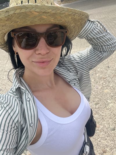 A woman, wearing a straw hat, sunglasses, and a striped button-up shirt over a white tank top, is taking a selfie outdoors. The background suggests an outdoor setting with gravel and a paved surface. The sunglasses reflect a scene with barrels and a person holding a phone, indicating the woman is taking the photo. The woman's hand is raised, adjusting her hat.