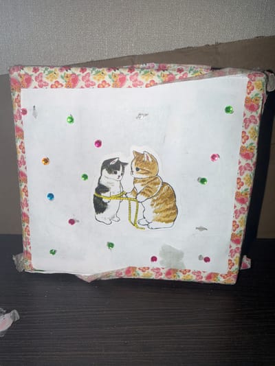 This is a close-up, eye-level shot of a handmade, decorated box. The box is covered in a floral and butterfly patterned fabric around the edges, with a white paper front. Two cartoon-style kitten stickers are affixed to the white paper; one is black and white, and the other is orange and white. They appear to be holding a yellow measuring tape between them. The box is adorned with numerous small, colorful sequins in red, green, blue, and orange scattered across the white front. The bottom of the box shows some wear and tear. The box rests on a dark, textured surface that appears to be a wooden table. The background is a textured, light-colored wall. The overall impression is of a whimsical, personalized craft item. There is no information available about its location in Zarech'e, Russia.