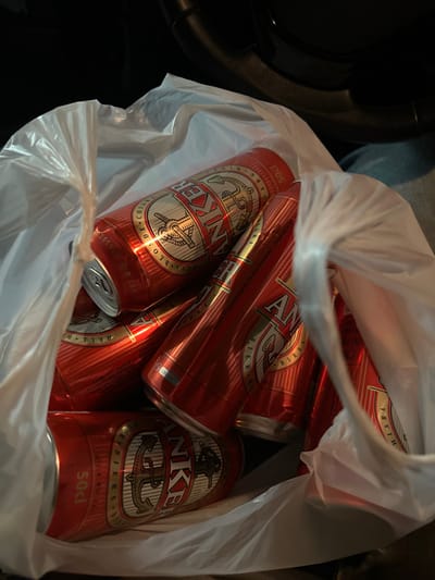 The image displays a white, translucent plastic shopping bag containing multiple red aluminum beverage cans. The cans are branded "ANKER" with a prominent anchor logo, text indicating "HELL BIER" and "BIRRA CHIARA," and a volume marking of "50cl." At least seven such cans are visible, stacked within the crumpled bag. The bag is positioned inside a vehicle, with a black steering wheel rim discernible in the upper background and a portion of what appears to be a person's leg clad in blue denim trousers visible on the right side of the frame. The lighting is dim, indicating an interior or nighttime setting. This scene is located in Wetzikon (ZH), Switzerland.