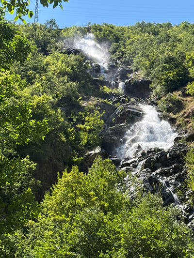 The image shows a cascading waterfall on a rocky, forested mountainside. Lush green trees and foliage cover much of the slope, with sunlight dappling through the leaves. The waterfall tumbles down over dark, irregular rocks, with spray creating a misty effect. In the background, a clear blue sky is visible, with power lines stretching across the top of the frame. The scene is natural and serene, with no people or man-made structures other than the power lines. The location is identified as the city of Pastra, Bulgaria, suggesting this is a natural landmark within or near the city.