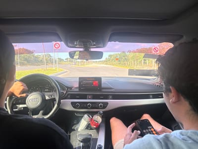 Inside an Audi, the driver is at the wheel, going 48 km/h, and navigating a turn on a sunny day. A passenger is on the phone, using the calculator app. Signs indicating no left turn and no entry are visible. An energy drink and a bottle of water sit in the central console of the car. Music is playing on the dashboard screen.