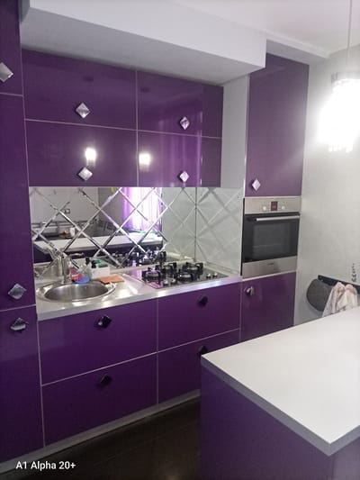 This media file depicts a modern kitchen with a bold, purple cabinetry scheme. The scene is well-lit, suggesting it's daytime or the kitchen is brightly illuminated. The subjects are the objects within the kitchen, specifically the sleek, high-gloss purple cabinets, a stainless steel sink with a modern faucet, a gas stovetop, and a built-in oven. A distinctive mirrored backsplash, made of square, beveled mirror tiles arranged in a diamond pattern, covers the wall behind the sink and stovetop, reflecting the surrounding kitchen elements. The cabinets are adorned with square, faceted drawer and door pulls that add a geometric accent.

To the right of the stovetop, a stainless steel built-in oven is visible, with a digital display showing "08:08". The oven is mounted within a tall purple cabinet. In the foreground, a portion of a white countertop extends into the frame, suggesting an island or a continuation of the counter space, also with purple cabinetry underneath.

The overall impression is one of a contemporary and stylish kitchen. There are no people visible, and the focus is entirely on the design and functionality of the space. The location context provided indicates this kitchen is located in Улукаўскі сельскі Савет, Belarus. The text "A1 Alpha 20+" is present in the bottom left corner of the image.