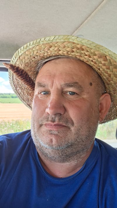 A close-up, eye-level, full face portrait of a middle-aged man wearing a straw hat and a blue t-shirt is shown. The man has light blue eyes, a mole on his left temple, and a grey beard. A feather is tucked into the brim of his hat. The background is blurred, showing a field and trees under a cloudy sky. The image appears to have been taken outdoors, possibly in a rural setting in Mereșeni, Moldova.