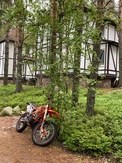 An orange dirt bike is parked on a dirt path next to a wooded area with a house in the background. The house is white with dark trim and a stone foundation. The dirt bike is in the foreground, leaning against a tree. The surrounding vegetation is green and lush. The sky is overcast. The location is Моторное, Russia.