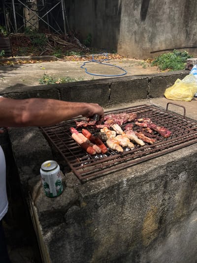 A person's arm, with visible sun spots, is reaching towards a grill laden with various meats, including sausages, chicken wings, and cuts of beef, cooking over an open flame. A can of beverage, featuring palm trees and Spanish text, sits on the concrete ledge beside the grill. In the background, the scene depicts an outdoor area with a concrete wall, dry leaves scattered on the ground, and what appears to be scaffolding or an industrial structure. The setting suggests a casual outdoor cooking event, possibly in a residential or communal outdoor space in San Juan del Sur, Nicaragua.