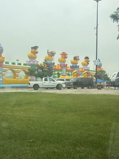 In Schaumburg, United States of America, an overcast day frames an outdoor scene dominated by a large, colorful inflatable playground resembling a castle. Various oversized inflatable ducks, each adorned with different accessories like hats, goggles, and sunglasses, are perched atop the inflatable structure, forming a whimsical skyline. In the foreground, a grassy area leads to a parking lot where several vehicles are parked, including a white pickup truck, a black minivan, and a recreational vehicle. A tall pole with security cameras and a sign that reads "GG 55" is visible, along with a small blue tent and some fencing further back. The overall atmosphere is one of a cheerful, albeit slightly muted, outdoor event.