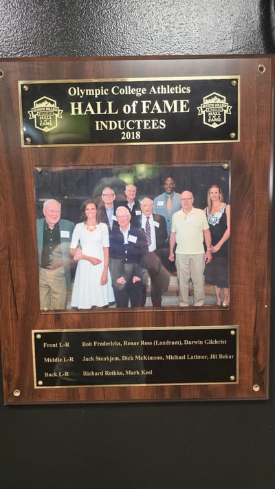 The image shows a framed plaque dedicated to the Olympic College Athletics Hall of Fame Inductees of 2018, located in Bremerton, United States of America. The plaque features a black background with gold lettering and a crest on the top left. Below the text, a photograph displays a group of nine individuals, presumably the inductees and possibly college representatives, posing for a picture. The photograph is protected by a clear plastic cover. Underneath the photograph, a black plaque with gold lettering lists the names of the inductees, organized by row: "Front L-R Bob Fredericks, Renae Ross (Landram), Darwin Gilchrist", "Middle L-R Jack Stenhjem, Dick McKimson, Michael Latimer, Jill Bekar", and "Back L-R Richard Rethke, Mark Keel". The plaque is mounted on a wooden board with a dark wood grain finish and is secured to a textured black surface in the background.