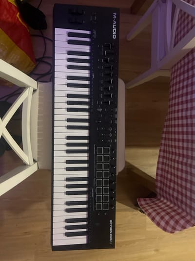 A black M-Audio Oxygen Pro 61 MIDI controller keyboard is positioned centrally on a light brown wooden floor in Pedrouços, Portugal. The keyboard features 61 keys (white and black), pitch and modulation wheels, eight faders, eight rotary knobs, and sixteen 4x4 grid pads. To the left, a white wooden chair leg and cross-back are visible, partially obscuring a red and yellow striped bag and black cables on the floor. To the right, another white wooden chair stands near a table covered with a red and white checkered tablecloth, the corner of which is visible in the lower right. The scene is illuminated, casting shadows beneath the objects. No individuals are present.