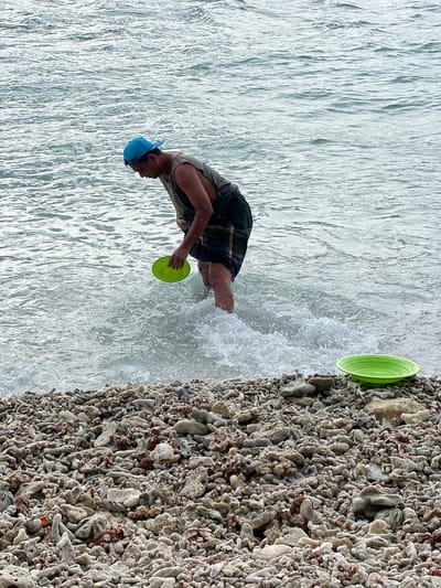 A man wearing a blue baseball cap, a sleeveless shirt, and dark patterned shorts stands in the shallow water at the edge of a coral-strewn beach. The man is holding a green disc in his right hand. Another green disc is placed on the beach to the right of the frame. The water is choppy and appears to be breaking around the man's legs. The beach is covered with various pieces of coral and rocks. The scene is located in Ujongsedheun, Indonesia.