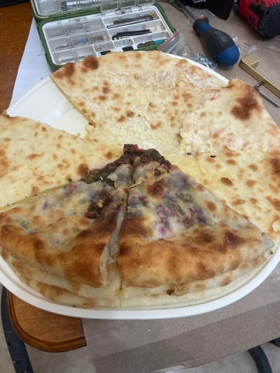 A close-up, overhead shot shows a round white plate filled with flatbreads cut into wedges. Some wedges are plain, golden-brown on the outside with a creamy filling, while others are filled with a dark green and purple mixture. The plate sits on a wooden surface next to a cardboard mat. In the background, a green toolbox filled with small tools, a blue-handled screwdriver, and a red and black power tool are visible. The scene appears to be an indoor setting, possibly a home or workshop, with a slightly cluttered arrangement of objects. The lighting is natural and diffused, casting soft shadows.