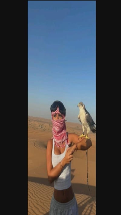 A woman stands in a desert landscape, likely Dubai, dressed in a white tank top and grey sweatpants. Her face is partially covered with a red and white checkered scarf, and goggles rest on her head. A falcon perches on her left arm, tethered with a cord. The background features rolling sand dunes and a clear blue sky.