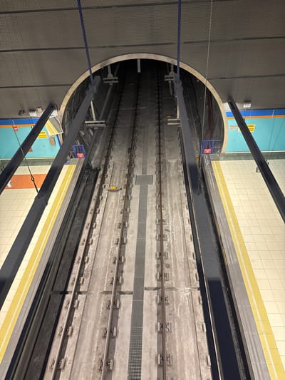 This is a high-angle, eye-level, full shot of a subway track in San Fernando de Henares, Spain. The image features two parallel subway tracks made of concrete and metal, disappearing into a large, circular tunnel. The tracks are lined with a yellow and grey platform, separated by a dark grey barrier. The ceiling is a dark grey, textured material, supported by dark grey beams. In the background, a blue wall with yellow and orange signage is visible. The scene appears to be an empty subway station, possibly under construction or maintenance, as there are no people or trains present. The lighting suggests it is daytime, with artificial lights illuminating the tracks and platform.