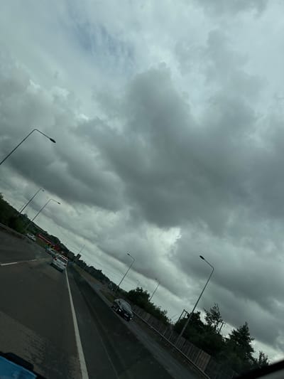 The image captures a view from a vehicle traveling on a highway, likely in or near Slieverue, Ireland, under a heavily overcast sky. The road surface appears wet, suggesting recent or ongoing precipitation. Several cars are visible on the multi-lane highway, indicating moderate traffic. Streetlights line the road at regular intervals, their lamps illuminated against the dim conditions. Vegetation lines the side of the highway, with trees and shrubs forming a green barrier. A large billboard with red text, possibly advertising "NolanTransport.com", is visible in the distance. The overall atmosphere is moody and somber due to the dark, cloudy sky, which dominates the upper portion of the frame. The perspective is angled, giving a dynamic feel to the scene of transit.