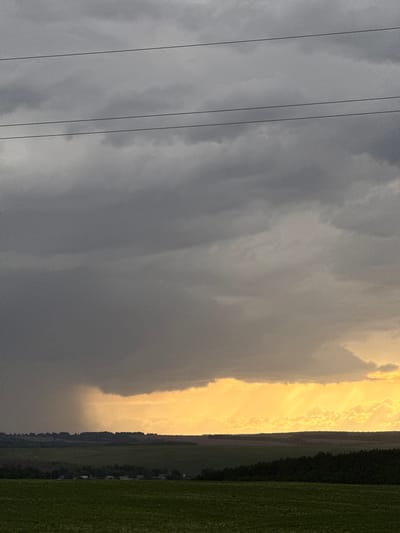 The image shows a landscape under a dramatic sky. In the foreground, power lines are visible against a backdrop of heavy, grey storm clouds. A contrasting patch of bright, yellow light illuminates a portion of the sky and possibly a downpour of rain, positioned over what appears to be a field. The right side of the image includes a dark treeline and a green field. Small structures, possibly buildings, are discernible near the treeline. There is no confirmed information about people being present in the image. The location in the city of Крынды, Russia, is also unconfirmed.