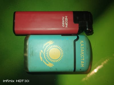 This is a top-down, close-up, slightly angled photograph of two lighters placed on a green, textured surface. The top object is a red disposable lighter with black accents, featuring the text "NEON TURBO" in white, vertically oriented text on its side. The bottom object is a light blue cigarette case with a black stripe along its right edge. It is adorned with a yellow emblem resembling the flag of Kazakhstan, which includes a sun and a steppe eagle. The text "ҚАЗАҚСТАН" (Kazakhstan in Kazakh) is also printed in yellow, vertically oriented on its right side. The lower left corner of the image displays the text "Infinix HOT 30i", indicating the device used to capture the photo. The surface appears to be a table or a similar flat area. The lighting suggests an indoor setting, with a bright glare reflecting on the green surface. There is no visible indication of the specific location within the city of Lisakovsk, Kazakhstan.