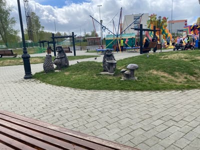 The image depicts a park in Kirovgrad, Russia. In the foreground is a wooden bench and a paved walkway. In the midground is a grassy knoll featuring decorative sculptures of hedgehogs and mushrooms. Behind the knoll are playground attractions including trampolines and inflatable structures. People, including children, are engaged in activities like jumping on trampolines and walking near the inflatable structures. A lamppost stands near the walkway. Trees are visible in the background, along with a lattice tower and buildings. The sky is cloudy.