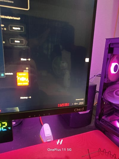 The image shows a computer setup in a room in Rajshahi, Bangladesh. The computer is displaying a black screen with a few icons and buttons. The computer is connected to a black box, possibly a subwoofer, and a large computer case with pink lighting. There are wires running from the computer case. A red mousepad is on the desk, and a white dongle, labeled "Satechi," is plugged in near the bottom of the image. The image is taken at a low angle, focused on the desk, and the computer screen is the focal point. The room appears dark, with a pink light emanating from the computer case. The photo is likely taken with a phone, indicated by the "H" and "OnePlus 11 5G" text in the lower right corner. The image depicts a typical setup for a computer user, with the screen displaying a blank screen and a few icons visible. The lighting and composition suggest that the image was captured in an indoor setting, likely a bedroom or home office. The specific details of the computer and other equipment provide context about the user's technological preferences.