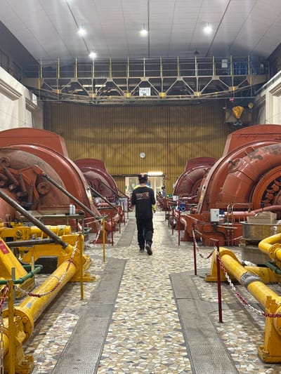 The image depicts the interior of a large industrial power generation facility in Pellafol, France. The scene features a long, high-ceilinged hall with two parallel rows of large, reddish-orange industrial machines, resembling turbines or generators, extending into the background. These machines display visible mechanical components, piping, and signs of wear and rust.

A central walkway separates the machinery rows. The floor of this aisle is composed of a multicolored mosaic tile pattern, interrupted by two parallel recessed metal grate sections running lengthwise. In the immediate foreground, yellow-painted heavy industrial equipment is present on both sides of the walkway, partially demarcated by red posts and white and red safety chains.

One individual, a male, is observed from behind, walking away from the viewer down the central aisle. He is wearing a dark, long-sleeved shirt with a yellow "ATOMS" logo on the back, dark trousers, and dark shoes. He is also wearing a yellow/orange hard hat or protective headwear.

Above the machinery, a prominent metal truss structure or catwalk with railings spans the width of the room, near the ceiling. A yellow sign on this structure reads "FORCE MAX 10T." Overhead crane hooks are visible towards the upper right of the image, associated with this structure. A circular white clock with black numerals is mounted on the far wall, centered above the walkway. The walls are light brown and appear to have a corrugated or ribbed texture. The ceiling is white and equipped with multiple overhead light fixtures.