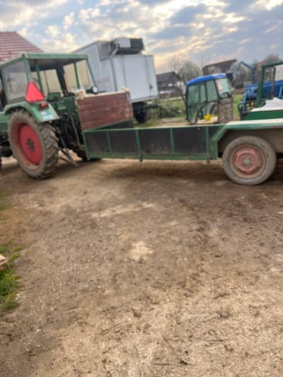 The image depicts an outdoor scene in what appears to be a rural or semi-rural setting within Krško, Slovenia. In the foreground, a dark green tractor with a red rear wheel hub is prominently featured on the left, hitched to a green, open-topped utility trailer. The tractor has a enclosed cab and a red triangular reflective sign mounted on its rear. A brown wooden crate rests between the tractor and the trailer's hitch point. The trailer, partially visible on the right, has a single red-hubbed wheel and black interior walls.
The ground is composed of compacted brown dirt and gravel, exhibiting tire tracks. In the mid-ground, behind the green tractor and trailer, a white refrigerated trailer or truck is parked. Further into the background, a detached blue and green tractor cab is resting on the ground, and another blue tractor is partially visible. Buildings with red roofs and sparse trees are present in the far background under a cloudy sky. No individuals are visible in the scene.