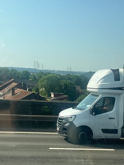 The image shows a view of the sky and a landscape from a moving vehicle. In the foreground, the side of a white van with a dark grey bumper and a raised, white cabin structure is visible, traveling on a road. The driver is partially visible in the driver's seat. The middle ground features a row of houses with reddish-brown roofs, and further beyond, a dense green forest. In the background, several tall, lattice-like electricity pylons are visible against a pale blue sky, suggesting a power grid in the distance. The lighting and hazy atmosphere indicate it is daytime, likely a sunny day. The scene appears to be a roadside view in or around Herstal, Belgium, given the presence of residential areas and significant infrastructure like power lines.
