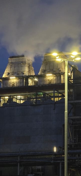 This image depicts an industrial facility in Frechen, Germany, at dusk or night. Two large, trapezoidal or conical-roofed industrial structures dominate the upper half of the frame, emitting significant plumes of white vapor or steam upwards into a dark blue sky. These structures feature multiple levels with horizontal rows of bright, yellowish-orange industrial lights illuminating their facades, platforms, and catwalks. In the foreground, positioned to the right, a tall metallic streetlamp with multiple radially arranged yellowish-orange luminaires stands upright, providing additional illumination to the scene. Below the main structures, a complex arrangement of darker concrete walls, metallic piping, and support structures is visible, also partially lit by the facility's ambient lighting. The overall environment indicates an active industrial operation. No individuals are present in the frame.