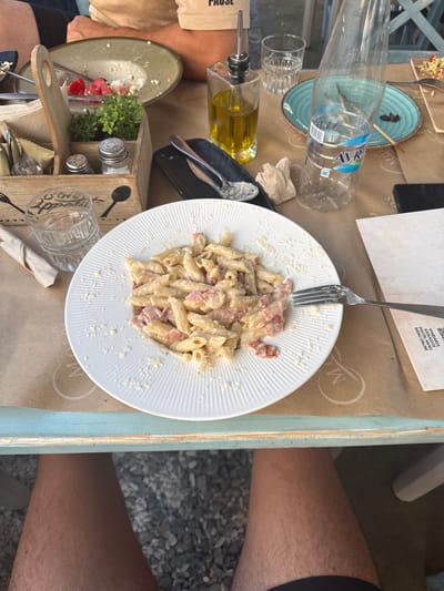 A high-angle, medium shot captures a table setting at a restaurant in Limenaria, Greece. A white plate of penne pasta with ham and cheese is centrally positioned on a brown paper tablecloth. To the left of the pasta, a wooden caddy holds salt and pepper shakers, napkins, and a small plant. A glass of water and a bottle of Avra brand mineral water are also present on the table. In the background, an empty salad plate and a bottle of olive oil are visible. A person's legs and lap are in the foreground, suggesting they are seated at the table. The outdoor setting is implied by the gravel on the ground visible beneath the table.