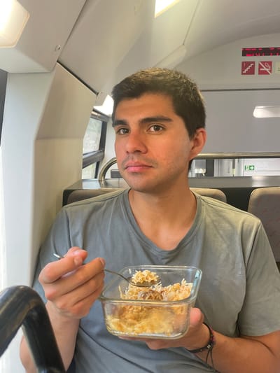 A young man with short, dark hair is seated on a train, holding a clear glass container filled with rice and a spoon. He is looking towards the camera with a neutral expression. The train interior features grey seats with red accents, a window with a view of greenery, and overhead compartments. Safety signs, including one depicting stairs and another prohibiting smoking, are visible. The scene appears to be in Gavà, Spain, suggested by the train context.