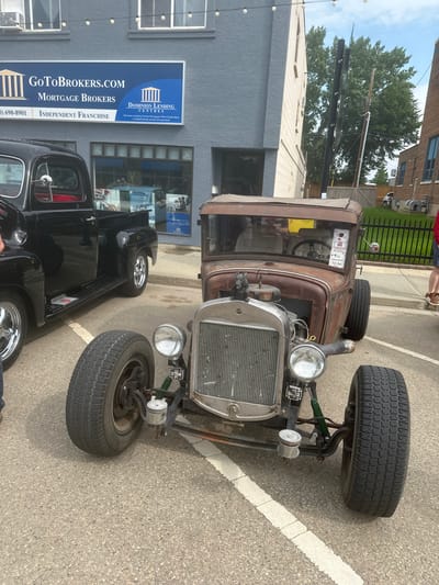 This is a vertical, eye-level shot of a rustic, rusty hot rod parked on an asphalt street. The hot rod, the primary subject, is a vintage vehicle with exposed components and a weathered exterior. Its large, knobby tires are prominently featured. To the left of the hot rod is a black pickup truck. Behind the vehicles is a building with a gray textured facade and large windows. A blue and white sign is attached to the building's exterior. The sign displays text that includes "GOTOBROKERS.COM", "MORTGAGE BROKERS", "DOMINION LENDING CENTRES", and a phone number.
The scene appears to be an outdoor event, possibly a car show or a street fair, given the presence of multiple vehicles and a commercial building. The lighting suggests it is daytime, likely with overcast or diffused sunlight as there are no strong shadows. The overall atmosphere is casual and relaxed, typical of a community gathering.
Visible text on the hot rod itself includes "#63" and "Rat Rod" on a small sign displayed on the windshield. The environment suggests an urban or suburban setting, possibly in the City of Leduc, Canada, as indicated by the location context. The focus is on the vintage vehicles and the commercial signage, creating a snapshot of a local event.