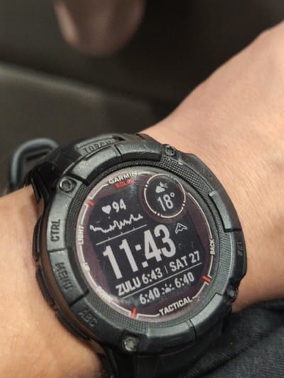 A black Garmin "TACTICAL" solar smartwatch is depicted on the left wrist of an individual. The watch face, illuminated and digital, displays a heart rate of 94 beats per minute, an ambient temperature of 18 degrees Celsius, and the current time as 11:43. Below the main time, "ZULU 6:43 | SAT 27" indicates Zulu time and the date. Further below, "6:40" is shown adjacent to both sunrise and sunset icons. The watch bezel and buttons are visible, with labels such as "GARMIN SOLAR," "TORCH," "CTRL," "MENU," "ABC," "LIGHT," "UP," "DOWN," "BACK," and "SET." The background is a dark, out-of-focus blur, offering no discernible details about the immediate environment or specific geographical features. The location within the city of San Bernardino, United States, cannot be determined from the provided image.