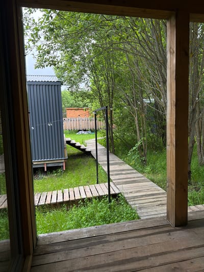 The image displays a lush outdoor scene viewed through a wooden window or door frame. The foreground consists of three sides of a light brown wooden frame, suggesting an interior perspective looking outward. The landscape beyond is dominated by abundant green foliage and trees, featuring numerous intertwining branches, some appearing bare or covered in lichen, and others densely packed with bright green leaves.

A rustic wooden boardwalk, constructed from parallel planks, meanders through the verdant ground, which is covered in green grass and wildflowers. This boardwalk includes a section with several steps, indicating a change in elevation. On the left side of the frame, a modern, dark grey corrugated metal structure, possibly a shed or a container, stands prominently. A string of decorative lights with clear, round bulbs is strung horizontally across the middle ground, adding an atmospheric element to the setting. In the distance, glimpses of another wooden fence or structure can be seen through the trees.

The lighting is soft and diffused, characteristic of an overcast day, and there are no visible harsh shadows. No people are present, and no text is visible. The scene evokes a tranquil, natural environment, possibly a garden, yard, or a retreat space.