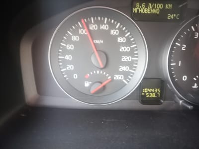 The image displays a car's dashboard with the speedometer showing approximately 100 km/h. To the upper right of the speedometer is a digital display showing "8.6 Л/100 KM МГНОВЕННО" which translates to "8.6 L/100 KM INSTANTANEOUS" and the ambient temperature as "24°C". Below this, a smaller digital display shows "104435 TI 538.7", likely indicating the odometer reading and a trip meter. The dashboard is lit, suggesting it is either nighttime or the car is in a dimly lit environment. The location is stated to be Perevolotsky, Russia.