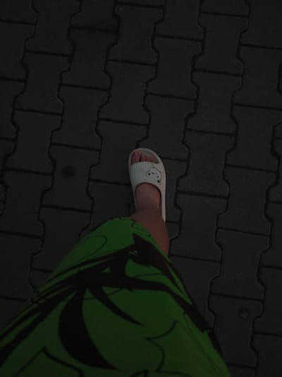 The image shows a person's foot wearing a white slide sandal with a cartoon bear design. The person is wearing a green garment with black patterns, possibly shorts or a skirt, which is partially visible in the lower left corner. The foot is positioned on a dark gray interlocking paver surface. The lighting suggests it is either early morning or late evening. The scene appears to be outdoors, on a paved walkway or street. There is no indication of the specific location within Galonki, Poland.