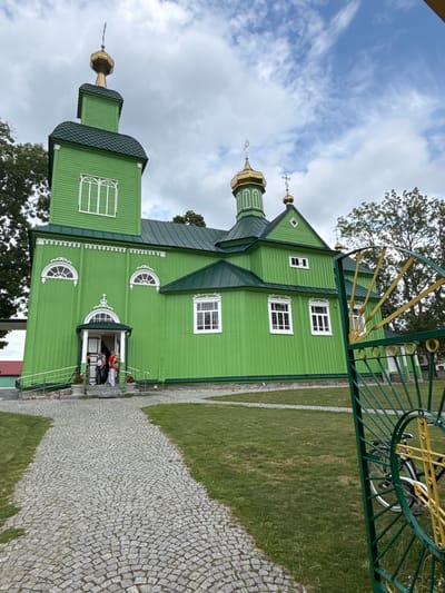In Trześcianka, Poland, a bright green wooden Orthodox church stands under a cloudy sky. The church features a tall, square bell tower topped with a golden dome and cross, as well as a larger, multi-gabled roof with several golden domes and crosses. The walls are clad in horizontal green wooden planks, and white-trimmed windows of various shapes and sizes punctuate the structure. An arched entryway with a small porch and white railings leads into the church, where several people are gathered. A cobblestone path leads to the entrance, flanked by a well-maintained lawn. In the foreground to the right, a green metal gate with yellow accents partially obscures a bicycle leaning against it. The surrounding environment consists of trees and grass.