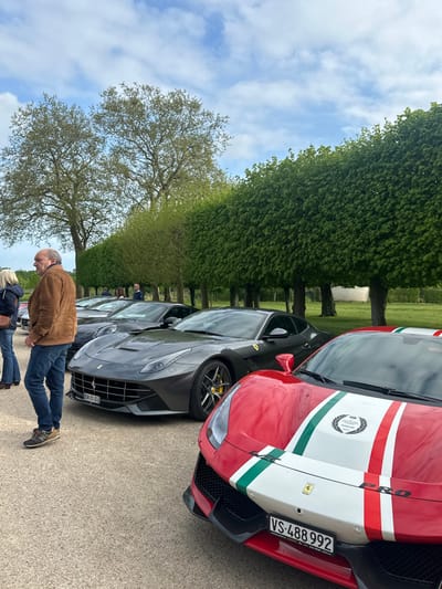 The scene takes place in the grounds of the Château de Chambord in the Loire Valley region of France, specifically in the landscaped gardens just west of the main chateau building. The image is captured from a ground-level perspective and showcases five individuals and four high-end sports cars.
Two individuals, a man and a woman, are standing in the foreground. To the left of the frame, a man in a brown jacket, blue jeans, and brown shoes stands with his back to the camera. He is engaged in a conversation with a woman in a dark jacket, her back turned to the camera. She is wearing blue jeans and black shoes.
There are two additional individuals visible in the background, also looking in the direction of the vehicles. They are positioned in the shade of the trees, their attire difficult to discern.
There are four sports cars present, all of the Ferrari brand. Two are positioned in the center of the frame. The closest to the camera is a red and white Ferrari with its hood up, showcasing a prominent Ferrari badge and a license plate reading "VS-488-992". It's followed by a dark grey Ferrari, its hood closed and its license plate obscured. Behind these two, a third dark grey Ferrari is partially visible, with its headlights obscured.
The vehicles are parked along a paved path, which disappears into the background amongst the trees and manicured gardens. In the background, a row of neatly trimmed trees creates a barrier between the path and the surrounding grounds. The trees, with their vibrant green foliage, are situated against a backdrop of a bright, cloud-filled sky.