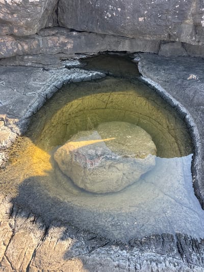 The image shows a close-up view of a natural rock formation containing a tide pool in Seoca, Montenegro. The rock is dark gray and textured with visible cracks and crevices. The tide pool is filled with clear water, reflecting the surrounding rocks and sunlight. A large, rounded rock rests within the tide pool, partially submerged. The water is shallow in some areas and deeper in others. The edges of the tide pool are irregular and show signs of erosion. Sunlight casts distinct shadows and illuminates parts of the water and rock, creating a contrast between light and dark areas. The environment appears to be coastal or near a body of water. No people or actions are depicted.