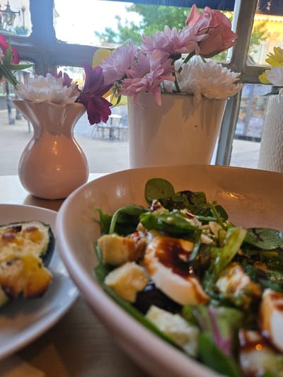 The image depicts a close-up view of a salad in a pale pink bowl, positioned on a wooden surface. The salad contains green leaves, white cheese cubes, and a dark drizzle, possibly balsamic glaze. To the left, a portion of another plate with what appears to be baked zucchini slices topped with cheese is visible. In the background, through a window, a blurred outdoor scene shows trees and building structures. Several vases filled with flowers are also present, with one pink vase containing white and purple flowers in the foreground, and another taller white vase with pink and white flowers further back. The overall setting suggests a dining environment, possibly a cafe or restaurant, with natural light coming from the window. There is no indication of the specific location in the Vale of White Horse, United Kingdom.