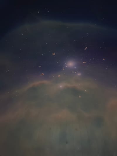 The image displays an interior view of a dark, domed projection space, likely a planetarium, located in Canela, Brazil. The environment is dimly lit, illuminated solely by the projected imagery. The central subject is a projection of a cosmic scene featuring a deep blue to purple background with numerous white and faint blue point light sources representing stars. Swirling, diffuse formations in shades of muted green, brown, and light orange, indicative of nebulae, are prominent across the mid-section. Bright, concentrated light clusters are visible within the projection. The lower portion of the image reveals the curved, paneled surface of the projection dome, characterized by faint vertical lines indicating structural seams. Scattered across the entire projected image are numerous irregularly shaped, opaque specks, varying in size, consistent with dust particles or condensation on a camera lens or the projection surface; some appear roundish, while others are elongated or tear-drop shaped. No individuals, specific objects beyond the projection and dome structure, or observable actions/interactions are present in the scene.