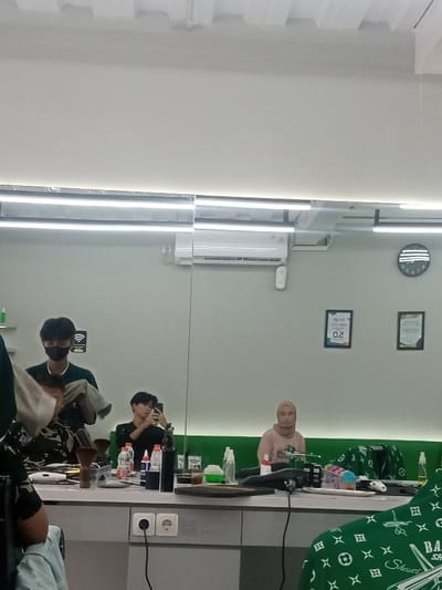 The image depicts the interior of a barber shop located in Sindanglaya, Indonesia. The scene is illuminated by overhead fluorescent strip lighting. A large mirror dominates the central and right portions of the frame, reflecting much of the shop's interior.
In the foreground, the back of a person's head and a green barber cape with white logos ("Barber Shaves" and scissors icon) are partially visible on the lower left. A grey counter with two electrical outlets is situated below this area.
Reflected in the mirror, from left to right:
1. A male barber, wearing a black face mask and a dark green shirt, stands, holding a white towel over the head of a client whose face is obscured.
2. A male client, seated in a barber chair, holds a black smartphone in his right hand, seemingly taking a photograph or video of himself. He wears a dark long-sleeved garment.
3. A female client, wearing a light brown hijab and a pink top, is seated on a green upholstered bench that runs along the wall. She faces forward.
The counter in front of the barber stations holds various items, including multiple spray bottles (some clear, some dark), plastic water bottles, and other hair products in dark containers. On the wall reflected in the mirror, an air conditioning unit is mounted above the counter. To the right of the air conditioner, a circular black wall clock with white markings is visible. Below the clock, two framed documents, one displaying "0.2," are mounted on the light-colored wall. A "WiFi" symbol is visible on a grey box on the wall to the left of the barber.