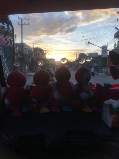 The image shows a view through a vehicle's window, likely a car or bus, in the urban area of RawangItiek, Indonesia. The window has several red plush toys attached with suction cups obscuring the view. Outside the window, the scene includes utility poles with wires, buildings with signage, and other vehicles. The sky is visible with a sunset or sunrise creating a bright glare. A person is walking on the street.