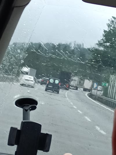 The image shows a view from inside a vehicle, looking out through a rain-streaked windshield onto a highway. The weather is overcast and rainy, indicated by the water droplets on the glass and the gray sky. Traffic is visible on the highway, with several cars and trucks in what appears to be a traffic jam.
In the foreground, a black phone mount with a suction cup is visible, attached to the windshield. Beyond the mount, the highway curves to the right. Cars and trucks are moving slowly or stopped on the road. A white car with a trailer is in the middle distance, and a black van is in front of it. Further down the road, more vehicles, including trucks, are visible. Trees line the side of the highway, suggesting a rural or suburban setting.
There are some signs visible on the right side of the highway, but they are too blurred to read. The overall mood is one of being stuck in traffic on a dreary, rainy day. There are no discernible people in the image, other than possibly hints of hands on the right edge of the frame. The time of day appears to be daytime, given the ambient light, but the heavy cloud cover makes it difficult to determine precisely. There is no visible text in the image.