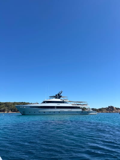 A large, white yacht is anchored in clear, blue water off the coast of a lush green island with rocky outcrops. The sky is a vibrant, cloudless blue. The yacht has multiple decks with awnings and railings. The island appears to be uninhabited, with dense vegetation. The scene suggests a calm, sunny day in a tropical or Mediterranean setting. Based on the provided context of La Maddalena, Italy, this image was likely captured in the archipelago, known for its pristine waters and numerous small islands.