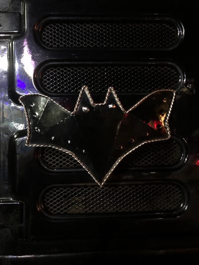 The image displays a close-up view of a black, glossy Batman emblem attached to a black, textured surface. The emblem, shaped like a bat with outstretched wings, features a metallic, braided border and several studs. The surface it is attached to has two horizontal, oval-shaped grilles, one above and one below the emblem, with a diamond pattern mesh. The lighting in the scene is dim, with reflections of colored lights, possibly purple and red, visible on the glossy surfaces. The background is dark and appears to be part of an interior structure. The image does not provide any context or information about the city of Karandugoda, Sri Lanka.
