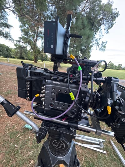 A Sony CineAlta camera sits mounted on a tripod outdoors. The camera has various cables connected, with one labeled "RB". A battery pack is attached to the camera's side. The tripod legs are metal, and the camera is pointed towards the sky with trees in the background. The ground beneath the tripod appears to be grassy.