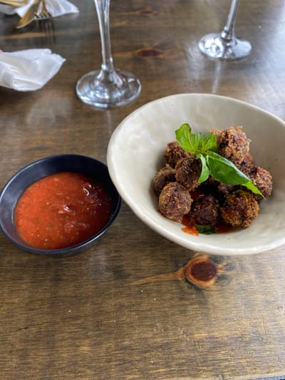 The scene depicts a close-up, elevated view of a dining table. In the foreground, a light beige, irregularly shaped ceramic bowl contains an arrangement of approximately 12-15 dark brown, breaded, spherical food items, possibly meatballs or croquettes, partially coated in a red-orange sauce. A sprig of fresh green basil rests on top of the food. To the left, a smaller, dark gray or black ceramic bowl holds a quantity of vibrant red-orange tomato-based sauce. Both bowls are positioned on a rustic, medium-brown wooden tabletop with visible grain and knots. In the blurred background, two empty stemmed wine glasses are visible, along with a folded white napkin and a portion of golden-colored cutlery (fork and knife) to the far left. No people are visible. The specific location within Puerto Almanza, Argentina, cannot be determined from the visual evidence provided.