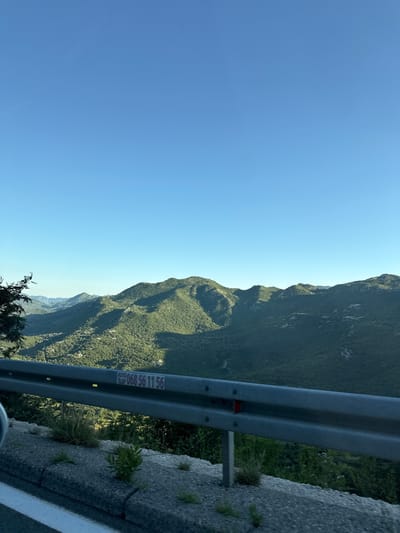 The image shows a view of forested mountains under a clear blue sky. In the foreground, a metal guardrail lines the side of a road. A concrete barrier with sparse vegetation is visible below the guardrail. A small sticker with the text "AUTO SLEP 068 56 11 56" is attached to the guardrail. The scene appears to be a scenic overlook, likely along a mountain road. The location is identified as Dobrsko selo, Montenegro. There are no people or specific actions depicted in the image.