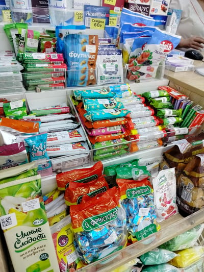 The image depicts a brightly lit shelf in a retail store, likely a pharmacy or convenience store, displaying a variety of packaged food items, predominantly sweets and snacks. The shelves are densely packed with colorful wrappers, featuring Russian text and branding. Prominently displayed are bags and boxes of "Фрутилал" (Frutilad) brand fruit snacks, including varieties like "Трио" (Trio) with nuts and dried fruits, and "Лесная ягода" (Forest Berry). Other items include "Нутринции" (Nutritions) berry-flavored gummies, "Умные Сладости" (Smart Sweets) jelly candies, and various individually wrapped candies. Some items are labeled with prices in Russian rubles and product descriptions such as "БЕЗ САХАРА" (sugar-free) and "С Фруктозой" (with fructose). In the background, a person's hand is visible reaching for an item, with a computer mouse on the counter. The overall impression is one of a well-stocked confectionery or health food section within a Russian retail establishment.